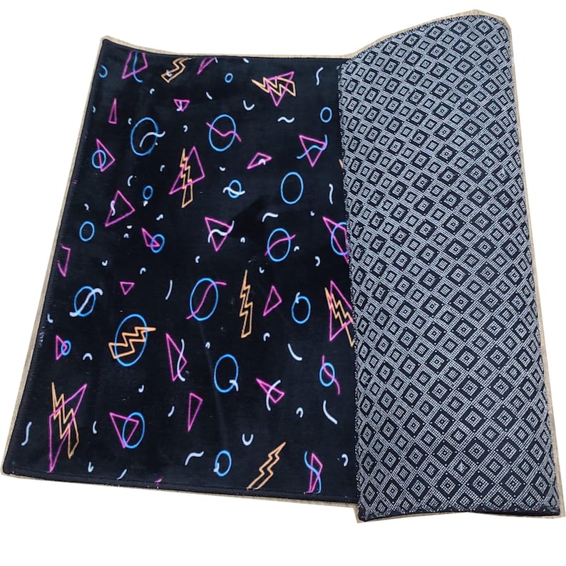 K&ouml;nnte beinhalten: Ein schwarzer rechteckiger Teppich mit einem Retro-Design der 80er Jahre. Der Teppich zeigt neonrosa, blaue und gelbe geometrische Formen, darunter Blitze, Kreise und Dreiecke. Der aufgerollte Abschnitt zeigt ein grau-wei&szlig;es Rautenmuster.