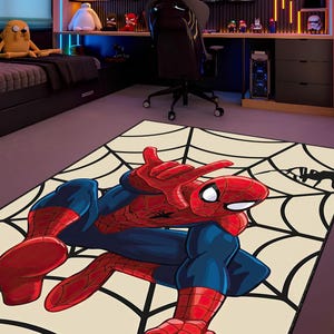 May include: A rectangular rug featuring a Spider-Man illustration in red and blue, posed on a cream-colored background with a black spiderweb design. The rug is in a room with a gaming setup and superhero figurines.