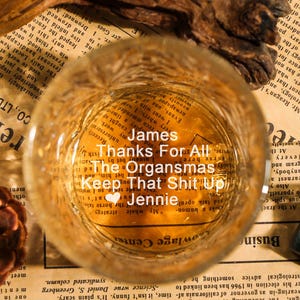 May include: A clear glass with text inside, resting on a newspaper. The text reads "James Thanks For All The Organsmas Keep That Shit Up Jennie". A pinecone and wood pieces are also present.