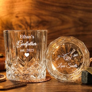 Puede incluir: Dos vasos de whisky transparentes grabados. Uno dice "Ethan's Godfather est. 2025" con un corazón. El otro vaso tiene un diseño de manos que se sostienen con el texto "Love Smith". Ambos vasos están sobre una superficie de madera.
