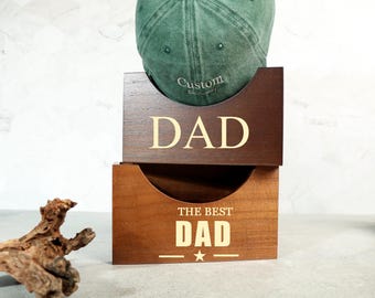 Puede incluir: Una gorra de béisbol verde descansa sobre dos cajas de madera. La caja superior está grabada con la palabra "DAD" en color claro. La caja inferior dice "THE BEST DAD" con una estrella. Las cajas son de color marrón medio.