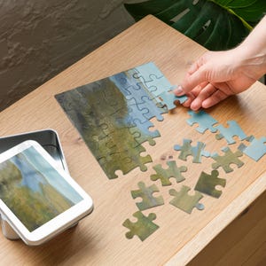 May include: A partially assembled jigsaw puzzle with a landscape painting design. The puzzle pieces are shades of green and blue. A hand is placing a light blue piece. A metal tin with the same image is on the left.