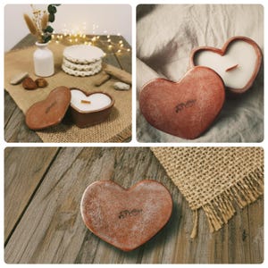 Puede incluir: Vela en forma de corazón en un recipiente de color terracota con cera blanca y una mecha de madera. El recipiente tiene una tapa con una inscripción. La vela se exhibe sobre una superficie de madera rústica.