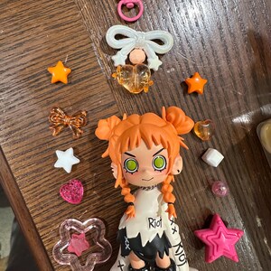 May include: A collection of colorful charms and a figurine on a wooden surface. The figurine has orange hair, green eyes, and black boots. The word "Riot" is printed on the figurine's shirt. Charms include stars, hearts, and bows in various colors.