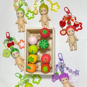 May include: A collection of small, nude-colored figurines with fruit-themed accessories. Each figure has a unique hairstyle and is paired with a keychain featuring beads and charms. A wooden crate holds miniature fruit.