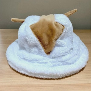 May include: A plush white bucket hat with a brown center and two small brown horns. The hat has a soft, fuzzy texture and a wide brim. The hat is designed to resemble a cow.