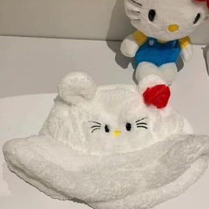 May include: A white plush bucket hat with a cat face design, featuring black whiskers, eyes, and a yellow nose. A red bow is on the side. A plush cat doll with a red bow, blue dungarees, and yellow top is in the background.