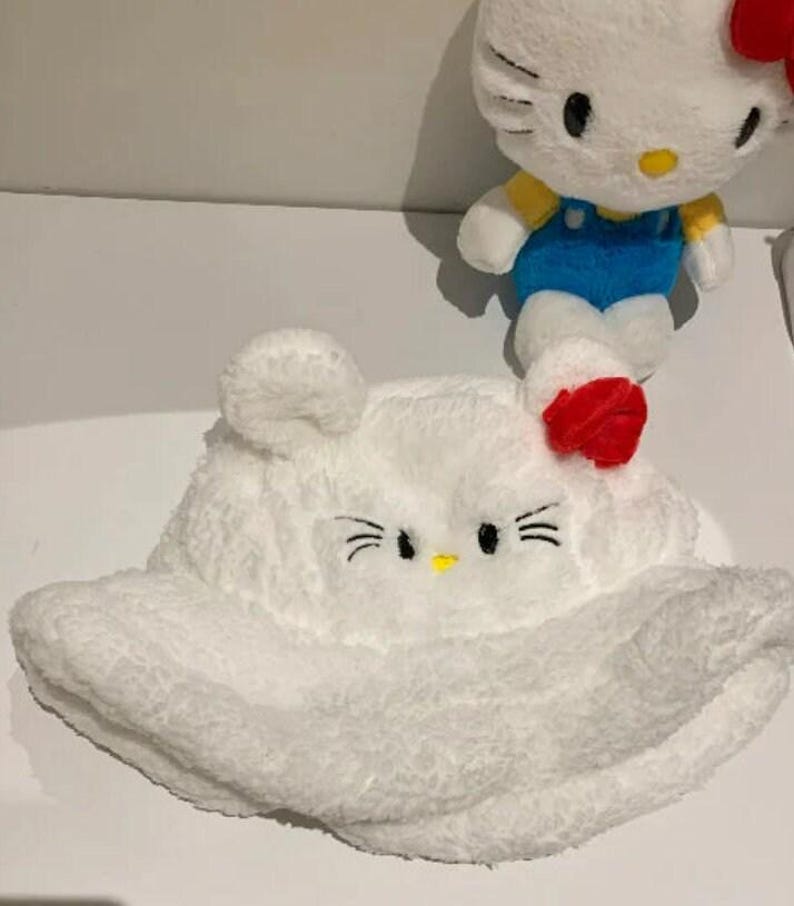 May include: A white plush bucket hat with a cat face design, featuring black whiskers, eyes, and a yellow nose. A red bow is on the side. A plush cat doll with a red bow, blue dungarees, and yellow top is in the background.