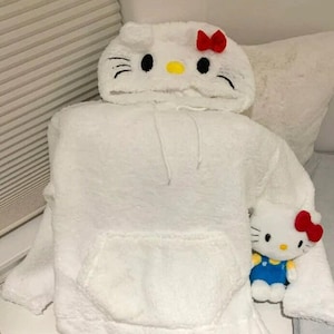 May include: A white, fluffy hoodie with a Hello Kitty design on the hood, featuring black eyes, whiskers, and a red bow. A matching Hello Kitty plush toy is also present, wearing blue overalls.
