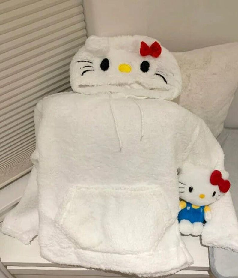 May include: A white, fluffy hoodie with a Hello Kitty design on the hood, featuring black eyes, whiskers, and a red bow. A matching Hello Kitty plush toy is also present, wearing blue overalls.