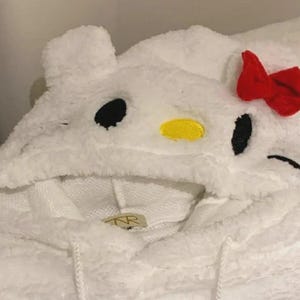 May include: White hooded sweatshirt with a cartoon character design. The hood features black eyes, a yellow nose, and a red bow. The sweatshirt is made of a soft, fuzzy material.