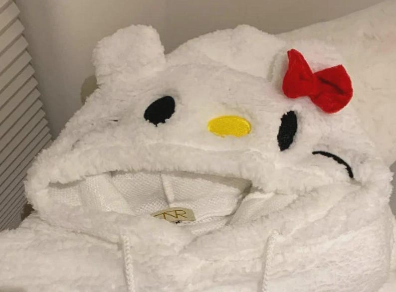 May include: White hooded sweatshirt with a cartoon character design. The hood features black eyes, a yellow nose, and a red bow. The sweatshirt is made of a soft, fuzzy material.