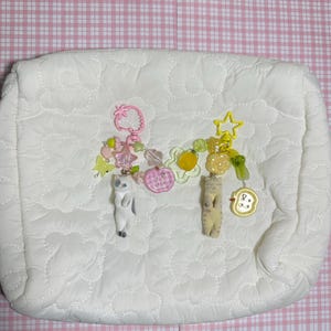 Puede incluir: Un neceser blanco acolchado con dos llaveros decorativos. Un llavero presenta una fresa, cuentas rosas y una figura de gato. El otro tiene una estrella amarilla, dijes de frutas y una figura de gato. El fondo es rosa y blanco.