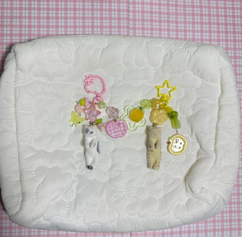 Puede incluir: Un neceser blanco acolchado con dos llaveros decorativos. Un llavero presenta una fresa, cuentas rosas y una figura de gato. El otro tiene una estrella amarilla, dijes de frutas y una figura de gato. El fondo es rosa y blanco.