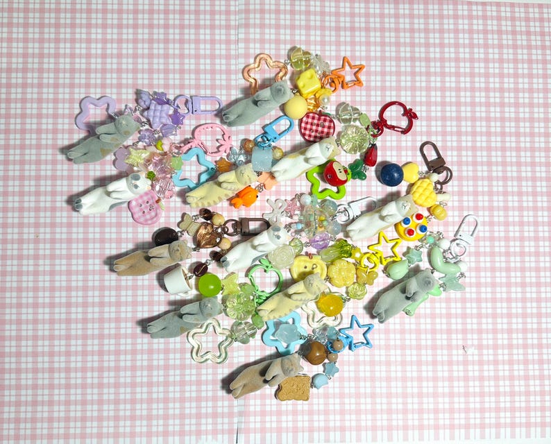 Puede incluir: Una colecci&oacute;n de llaveros y dijes coloridos, que incluyen figuras en forma de oso en varios colores, estrellas y otros elementos decorativos. Los art&iacute;culos est&aacute;n dispersos sobre una superficie a cuadros rosa y blanco.