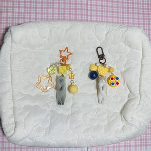 Puede incluir: Dos llaveros con dijes de gato y cuentas decorativas. Un llavero presenta un dije de gato gris, una estrella amarilla y una esfera amarilla. El otro llavero tiene un dije de gato blanco, cuentas de coraz&oacute;n amarillas y una esfera colorida con puntos azules y rojos.