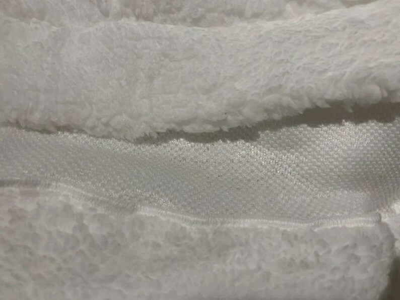 May include: Close-up of white fabric showing a contrast of textures. The image features a fluffy, plush material alongside a smooth, woven fabric. The textures create a layered effect, with the fluffy material on top.