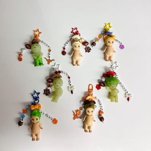 May include: A collection of small, nude-colored figurines, each adorned with a unique hat and accessories, including footballs and star-shaped charms. Some figurines have text like "HOOKS" and "GATORS" on beaded strings. The figurines are attached to keychains.