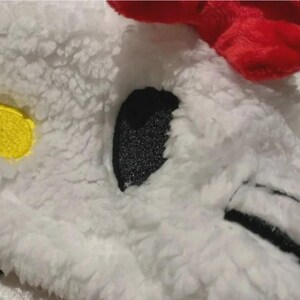 May include: Close-up of a white, fluffy fabric with a red accent. Features a black, glittery eye, a yellow patch, and black stitching. The texture suggests a soft, plush toy or accessory.