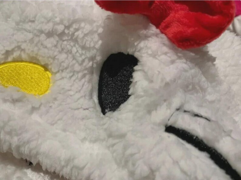 May include: Close-up of a white, fluffy fabric with a red accent. Features a black, glittery eye, a yellow patch, and black stitching. The texture suggests a soft, plush toy or accessory.