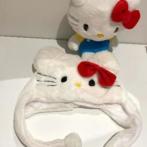 May include: A white plush Hello Kitty hat with ear flaps and a red bow, next to a Hello Kitty plush toy with a red bow and blue dungarees. The hat has black eyes and whiskers.
