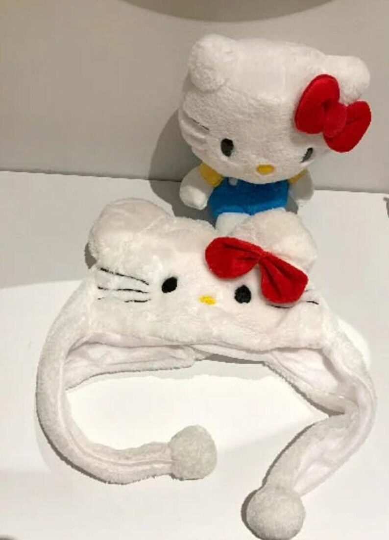 May include: A white plush Hello Kitty hat with ear flaps and a red bow, next to a Hello Kitty plush toy with a red bow and blue dungarees. The hat has black eyes and whiskers.