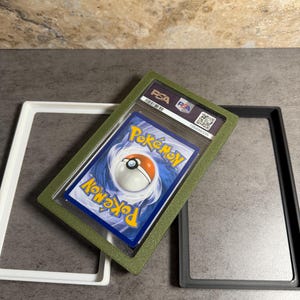 Op de afbeelding: Een beoordeelde Pok&eacute;mon-verzamelkaart in een groene beschermhoes, met het PSA-logo zichtbaar. De kaart wordt weergegeven met twee lege frames, een witte en een grijze. De kaart heeft een blauwe achtergrond met een Pok&eacute; Ball.