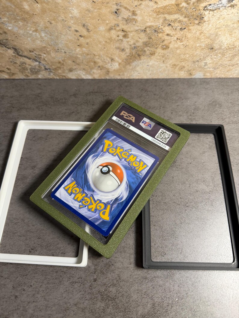 Op de afbeelding: Een beoordeelde Pok&eacute;mon-verzamelkaart in een groene beschermhoes, met het PSA-logo zichtbaar. De kaart wordt weergegeven met twee lege frames, een witte en een grijze. De kaart heeft een blauwe achtergrond met een Pok&eacute; Ball.