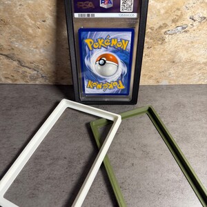 Op de afbeelding: Een beoordeelde Pok&eacute;mon-verzamelkaart in een zwarte beschermhoes. De kaart heeft een blauwe achtergrond met het Pok&eacute;mon-logo en een Pok&eacute; Ball-ontwerp. Twee rechthoekige frames, &eacute;&eacute;n wit en &eacute;&eacute;n olijfgroen, zijn ook zichtbaar.