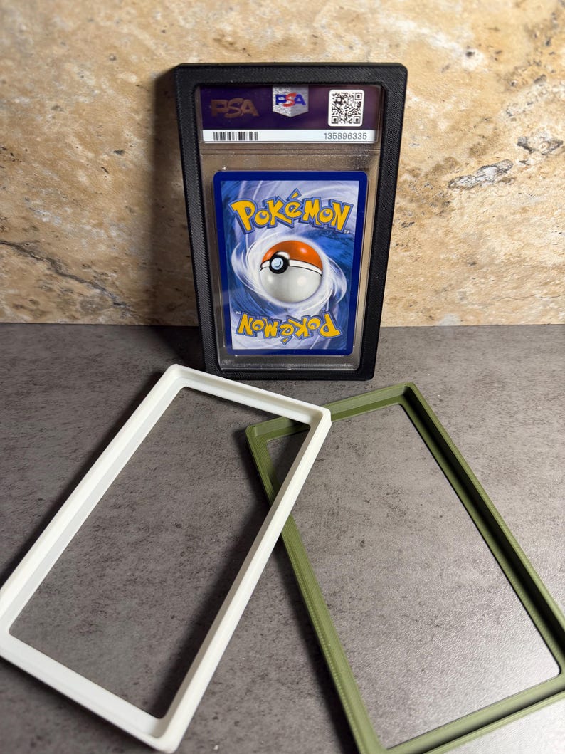 Op de afbeelding: Een beoordeelde Pok&eacute;mon-verzamelkaart in een zwarte beschermhoes. De kaart heeft een blauwe achtergrond met het Pok&eacute;mon-logo en een Pok&eacute; Ball-ontwerp. Twee rechthoekige frames, &eacute;&eacute;n wit en &eacute;&eacute;n olijfgroen, zijn ook zichtbaar.