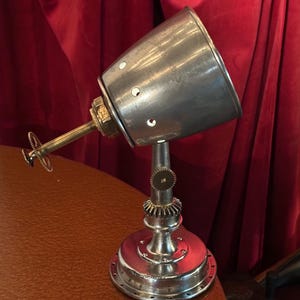 May include: A steampunk-style desk lamp with a metallic finish. The lamp features a conical shade with circular cutouts, a brass arm, and a base with gear-like details. It is positioned on a brown surface against a red backdrop.