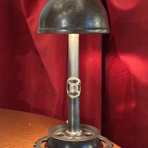 Puede incluir: Lámpara de mesa de estilo industrial. Presenta una pantalla oscura en forma de cúpula, una columna central de metal con un engranaje decorativo y una base circular con un borde en forma de engranaje. La lámpara es de metal y tiene una estética vintage.