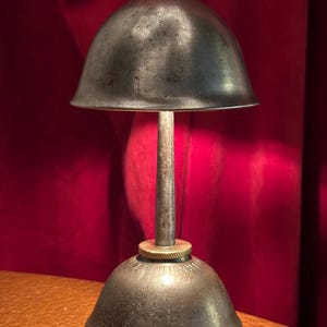 May include: A vintage metal oil can with a bell-shaped top and base, connected by a slender stem. The can has a weathered, aged appearance with brass accents. The background is a deep red curtain.