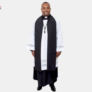May include: A person wearing a white clergy robe with black accents, including a black stole and a black collar. The robe has decorative trim on the sleeves. The Clergy Attire logo is in the upper left corner.