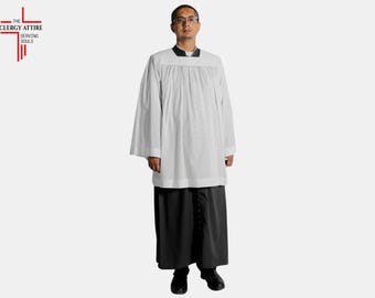 May include: A white clergy alb with wide sleeves and a black cassock. The alb has a gathered neckline and is worn with a black cassock. The Clergy Attire logo is in the upper left corner.