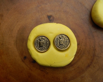 May include: Two antique brass-colored buttons with owl designs are set into a yellow, circular shape. The buttons are detailed with an owl surrounded by a wreath. The background is a dark brown wooden surface.