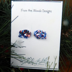 May include: A pair of stud earrings shaped like bison, featuring a vibrant mix of blue, red, and silver glitter. The earrings are mounted on a white display card printed with the text From the Woods Designs and a floral line art illustration.