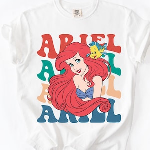 Op de afbeelding: Wit T-shirt met een kleurrijke afbeelding van Ariel uit De Kleine Zeemeermin. Het ontwerp bevat Ariel's afbeelding, een gele vis en de naam "ARIEL" in gelaagde, retro-stijl letters. Een leuke, casual tee.