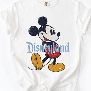 Puede incluir: Camiseta blanca con una ilustraci&oacute;n vintage de Mickey Mouse. Mickey aparece con pantalones cortos rojos y zapatos amarillos, con la palabra "Disneyland" en azul. El dise&ntilde;o tiene un aspecto envejecido y desgastado.