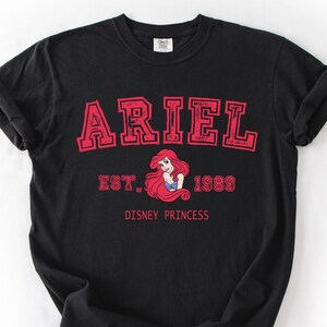 K&ouml;nnte beinhalten: Schwarzes T-Shirt mit dem Namen "ARIEL" in roter Schrift im College-Stil. Es enth&auml;lt "EST. 1989" und eine Grafik von Arielle aus Disneys "Arielle, die Meerjungfrau". Darunter steht "DISNEY PRINCESS".