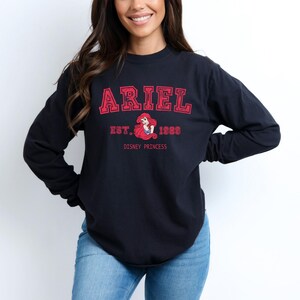 K&ouml;nnte beinhalten: Schwarzes Langarmshirt mit dem Namen "ARIEL" in roten Gro&szlig;buchstaben. Darunter steht "EST. 1989" und eine Grafik von Arielle, der Disney-Prinzessin. Die Worte "DISNEY PRINCESS" sind darunter gedruckt.