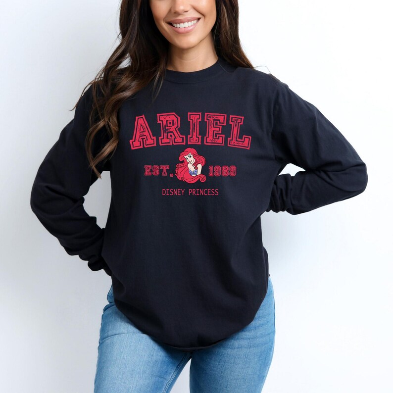 K&ouml;nnte beinhalten: Schwarzes Langarmshirt mit dem Namen "ARIEL" in roten Gro&szlig;buchstaben. Darunter steht "EST. 1989" und eine Grafik von Arielle, der Disney-Prinzessin. Die Worte "DISNEY PRINCESS" sind darunter gedruckt.