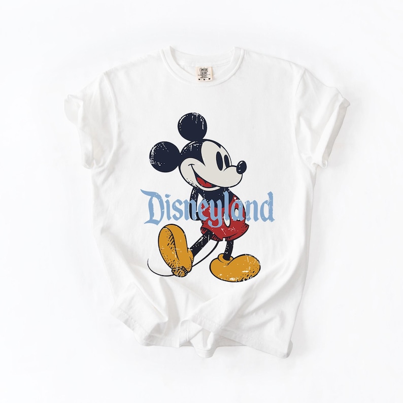 Puede incluir: Camiseta blanca con una ilustraci&oacute;n vintage de Mickey Mouse. Mickey aparece con pantalones cortos rojos y zapatos amarillos, con la palabra "Disneyland" en azul. El dise&ntilde;o tiene un aspecto envejecido y desgastado.