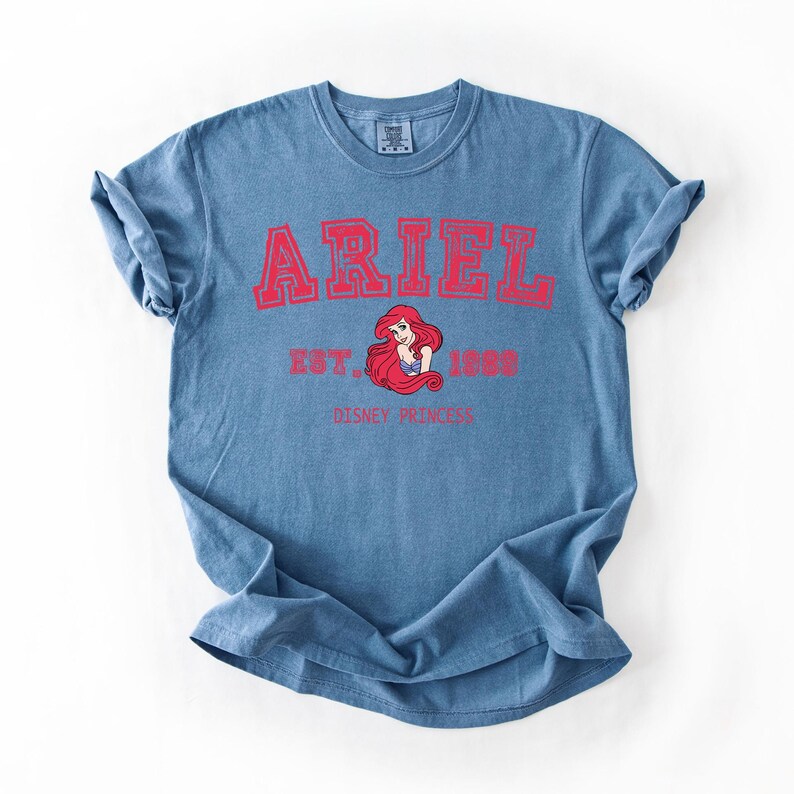 K&ouml;nnte beinhalten: Blaues T-Shirt mit dem Namen "ARIEL" in roten Blockbuchstaben. Darunter eine Grafik von Ariel, der Disney-Prinzessin, mit dem Text "EST. 1988" und "DISNEY PRINCESS" in Rot. Das T-Shirt ist gefaltet.