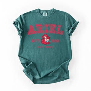 K&ouml;nnte beinhalten: T&uuml;rkisfarbenes T-Shirt mit dem Namen "ARIEL" in roten Blockbuchstaben. Darunter steht "EST. 1989" und eine Grafik von Arielle, der Disney-Prinzessin. Die Worte "DISNEY PRINCESS" sind unter der Grafik aufgedruckt.