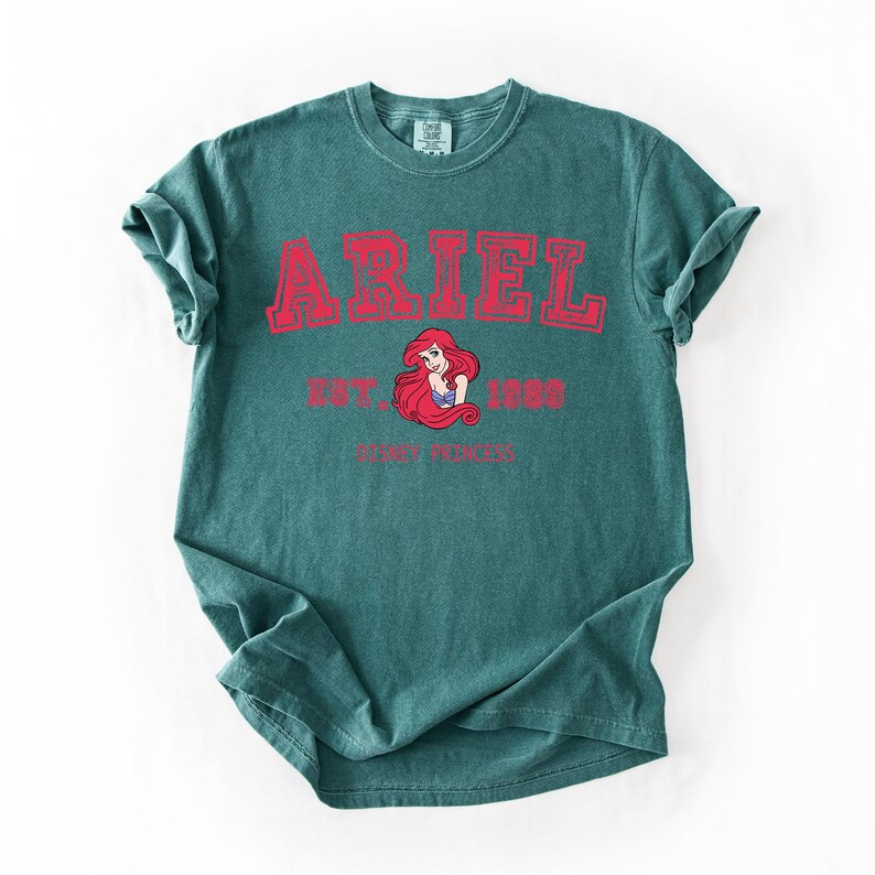 K&ouml;nnte beinhalten: T&uuml;rkisfarbenes T-Shirt mit dem Namen "ARIEL" in roten Blockbuchstaben. Darunter steht "EST. 1989" und eine Grafik von Arielle, der Disney-Prinzessin. Die Worte "DISNEY PRINCESS" sind unter der Grafik aufgedruckt.