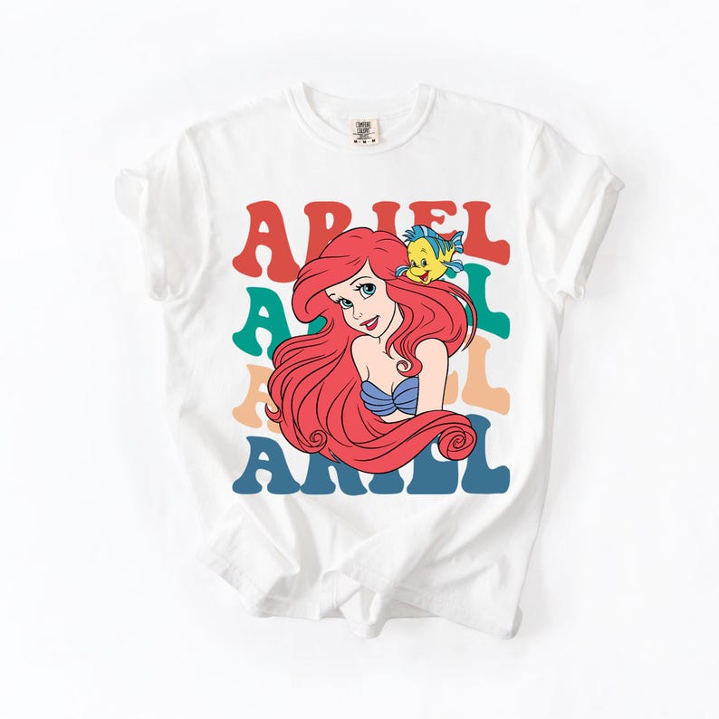 Op de afbeelding: Wit T-shirt met een kleurrijke afbeelding van Ariel uit De Kleine Zeemeermin. Het ontwerp bevat Ariel's afbeelding, een gele vis en de naam "ARIEL" in gelaagde, retro-stijl letters. Een leuke, casual tee.
