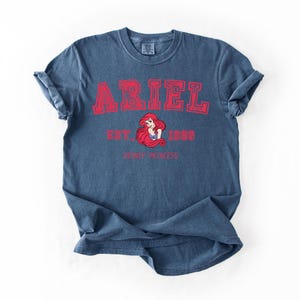 K&ouml;nnte beinhalten: Blaues T-Shirt mit dem Namen "ARIEL" in roten Blockbuchstaben. Darunter eine Grafik von Arielle, der Meerjungfrau von Disney, mit den Worten "EST. 1989" und "DISNEY PRINCESS" in Rot.