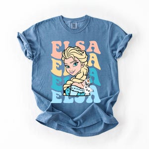 Puede incluir: Camiseta azul con un dise&ntilde;o de Elsa de Frozen, con el nombre "Elsa" en letras retro coloridas. Elsa se representa con cabello rubio y un vestido azul, junto a Olaf el mu&ntilde;eco de nieve.