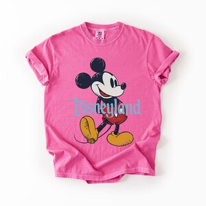 Puede incluir: Una camiseta rosa con un gr&aacute;fico de Mickey Mouse caminando y la palabra "Disneyland" en azul. Mickey Mouse es negro, blanco, rojo y amarillo. La camiseta tiene las mangas enrolladas.